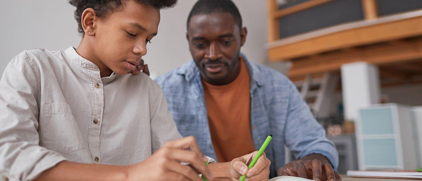 A parent looking over his son's shoulder as he writes in a notebook.